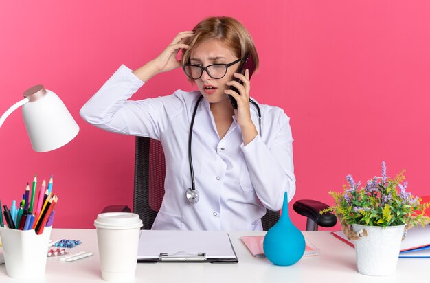 Besorgte junge ärztin, die ein medizinisches gewand mit stethoskop und brille trägt, sitzt am schreibtisch mit medizinischen werkzeugen spricht am telefon und legt die hand auf den kopf, isoliert auf rosa wand