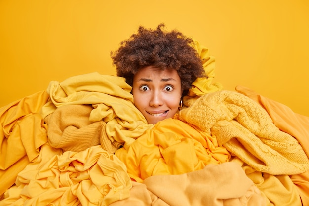 Besorgte junge Afro-Amerikanerin beißt Lippen, umgeben von gelber Kleidung, sammelt Gegenstände aus dem Kleiderschrank, um sie zu recyceln, steckt den Kopf durch die Kleidung und nimmt alles heraus