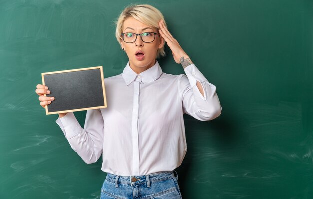 Besorgte junge blonde Lehrerin mit Brille im Klassenzimmer, die vor der Tafel steht und eine Mini-Tafel zeigt, die die Hand auf dem Kopf hält und mit Kopienraum nach vorne schaut