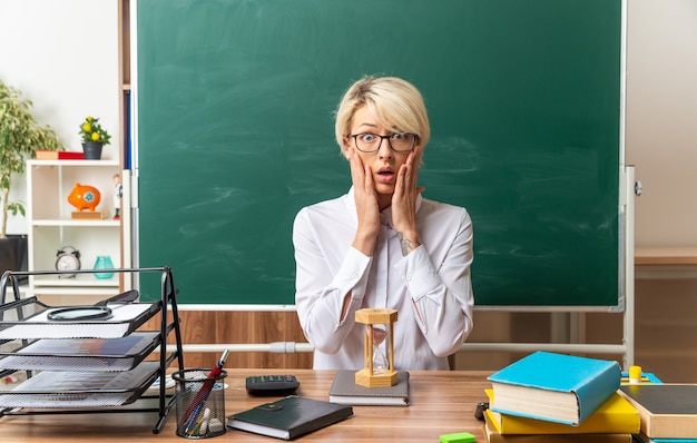 Besorgte junge blonde Lehrerin mit Brille sitzt am Schreibtisch mit Schulwerkzeugen im Klassenzimmer und hält die Hände auf dem Gesicht