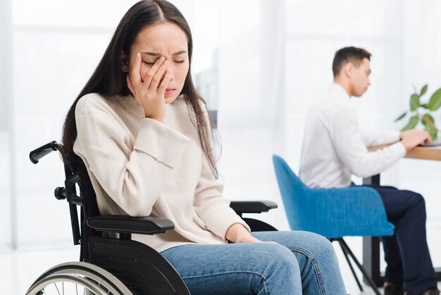 Besorgte junge Frau, die auf dem Rollstuhl sitzt vor dem männlichen Kollegen sitzt, der Laptop verwendet