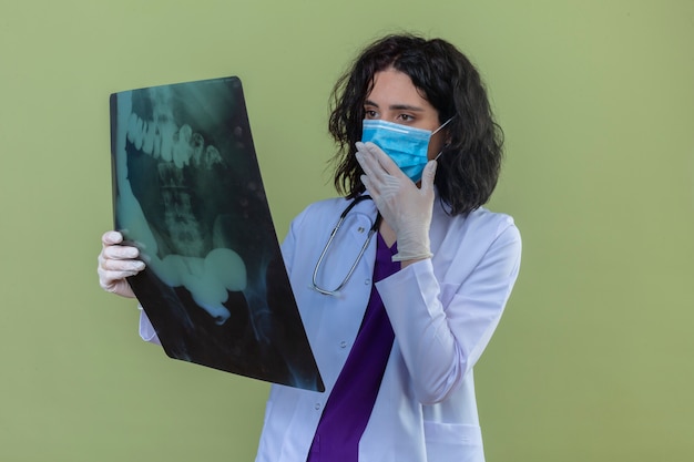 Besorgte junge Ärztin, die weißen Kittel mit Stethoskop in der medizinischen Schutzmaske trägt, die nervös auf Röntgenaufnahme der Lungen steht, die auf lokalisiertem Grün stehen