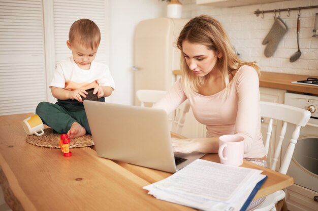 Besorgte verärgerte junge blonde Frau, die am Küchentisch mit Papieren und tragbarem Computer sitzt und sich gestresst fühlt, weil sie Bericht erstatten und auf ihren kleinen Sohn aufpassen muss, während er zu Hause bleibt