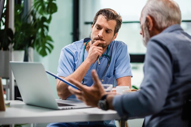 Besorgter Arzt, der medizinische Berichte seines Patienten auf einem Computer liest und während des Arzttermins über die Art der Behandlung nachdenkt