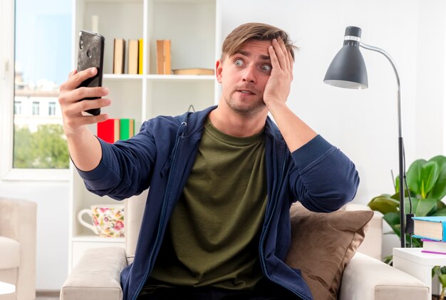 Besorgter junger blonder gutaussehender Mann sitzt auf einem Sessel und schaut auf das Telefon, das die Hand legt