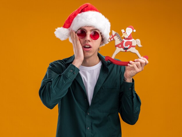 Besorgter junger blonder Mann mit Weihnachtsmütze und Brille, der den Weihnachtsmann auf einer Schaukelpferdfigur hält und in die Kamera schaut, die Hand auf dem Gesicht isoliert auf orangefarbenem Hintergrund hält keeping