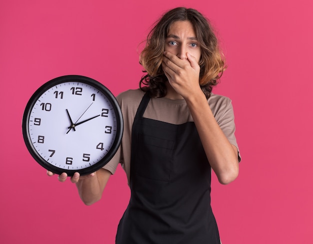 Besorgter junger, gutaussehender Friseur, der eine einheitliche Uhr trägt, die auf die Vorderseite schaut und die Hand auf den Mund legt, isoliert auf rosa Wand