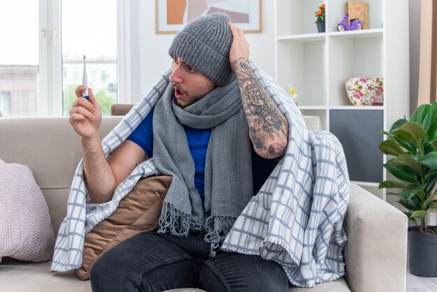 Besorgter junger kranker Mann mit Schal und Wintermütze, der in Decke gewickelt auf dem Sofa im Wohnzimmer sitzt und das Thermometer hält und die Hand auf dem Kopf hält