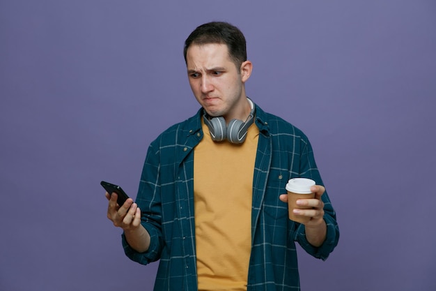 Besorgter junger männlicher Student mit Kopfhörern um den Hals, der eine Kaffeetasse aus Papier und ein Handy hält und das Handy isoliert auf violettem Hintergrund betrachtet