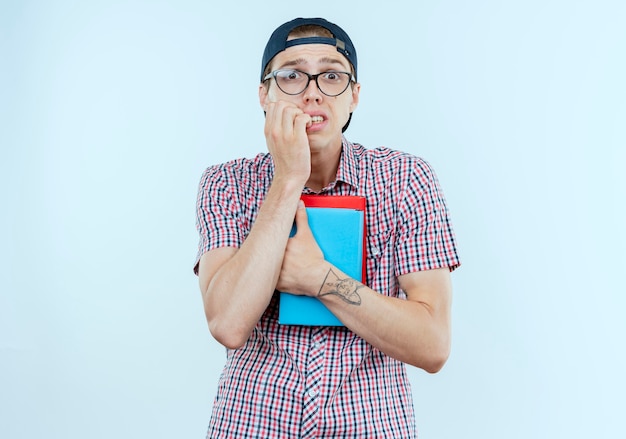 besorgter junger Student, der Rückentasche und Brille und Kappe hält, Bücher hält und Nägel beißt