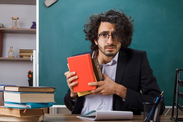 Besorgter männlicher Lehrer mit Brille, der ein Buch hält, das am Tisch mit Schulwerkzeugen im Klassenzimmer sitzt