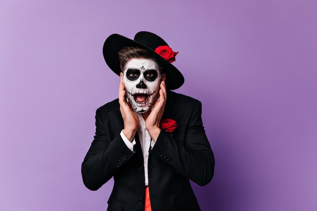 Besorgter Mann im traditionellen mexikanischen Kostüm, das am Tag der Toten aufwirft. Studioporträt des überraschten Zombietyps im schwarzen Hut.