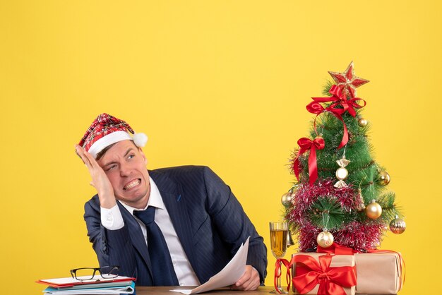 besorgter Mann mit Weihnachtsmütze, der am Tisch nahe Weihnachtsbaum sitzt und auf Gelb präsentiert