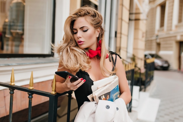Besorgtes blondes weibliches Modell, das soziale Netzwerke überprüft, während es neben Restaurant allein steht