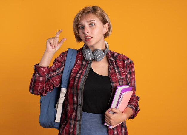 Besorgtes junges slawisches Studentenmädchen mit Kopfhörern, das Rucksack trägt, hält Buch und Notizbuch