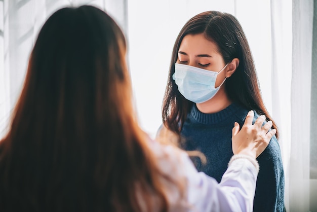 Besuch eines asiatischen Arztes und Untersuchung einer jungen erwachsenen Frau im Krankenhaus mit Bürosyndrom und Depression Der Arzt untersucht und konsultiert die Gesundheitsversorgung mit einer Maske zum Schutz der Covid19-Pandemie