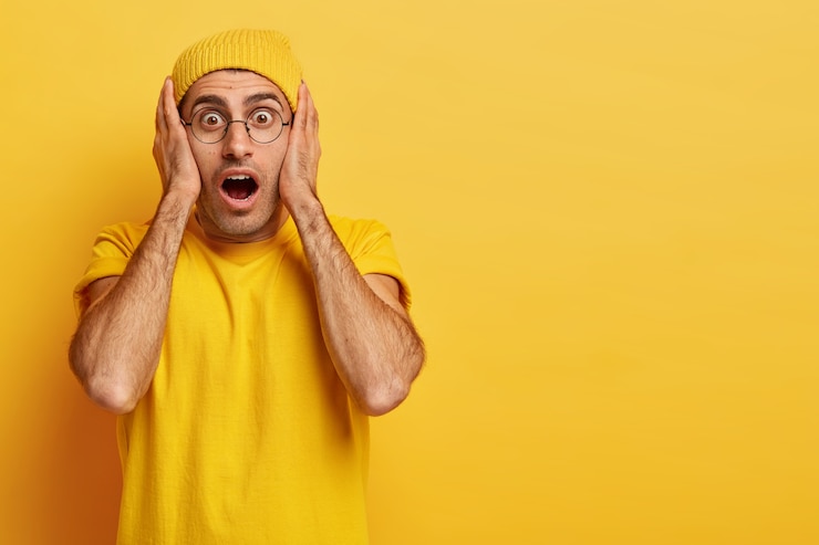 Kostenlose Foto betäubter emotionaler europäischer mann hält die hände im gesicht, den mund weit geöffnet, kann nicht an schockierende relevanz glauben, trägt leuchtend gelbe kleidung und ist sehr emotional. omg-konzept