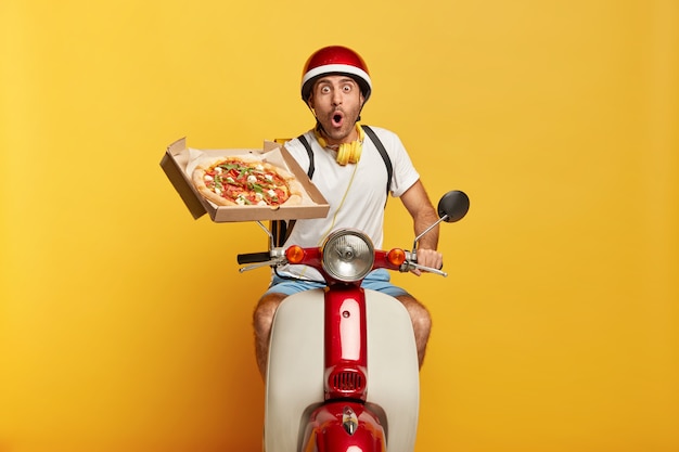 Betäubter hübscher männlicher Fahrer auf Roller mit rotem Helm, der Pizza liefert