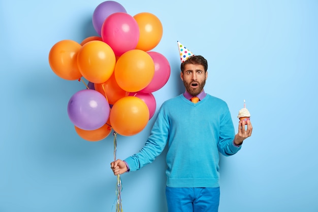 Betäubter Kerl mit Geburtstagshut und Luftballons, die im blauen Pullover aufwerfen