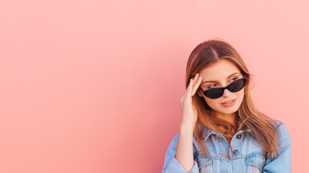 Betonen Sie die tragende Sonnenbrille der jungen Frau, die weg gegen Pfirsichfarbhintergrund schaut