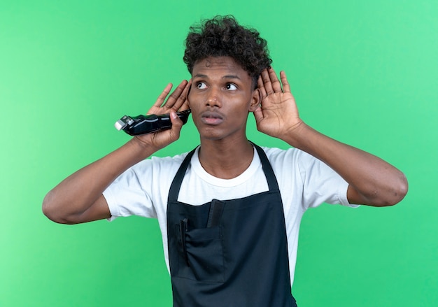 Betrachten der Seite denkend junger afroamerikanischer männlicher Friseur, der Uniform hält, die Haarschneidemaschinen hält und hörende Geste lokalisiert auf grüner Wand zeigt