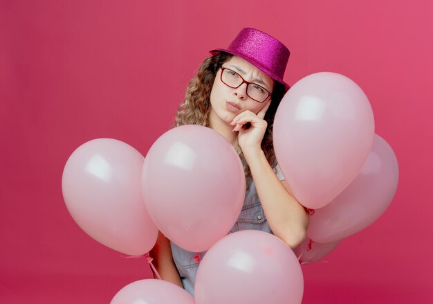 Betrachten der Seite denkendes junges Mädchen, das Brille und rosa Hut hält Ballons hält und Hand auf Wange lokalisiert auf rosa Hintergrund setzt