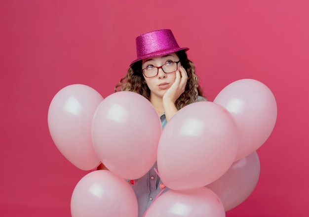 Betrachten der Seite denkendes junges Mädchen, das Brille und rosa Hut trägt, der unter Luftballons steht und Hand auf Wange lokalisiert auf rosa Hintergrund setzt