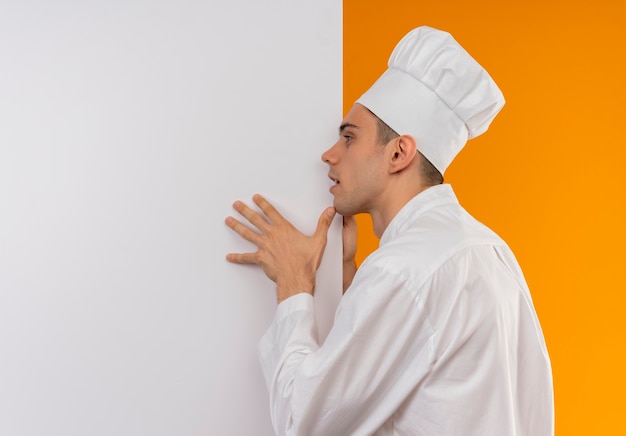 Betrachten der Seite junger männlicher kühler tragender Kochuniform, die weiße Wand auf isolierter gelber Wand mit Kopienraum hält