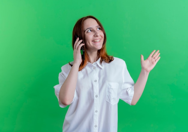 Betrachten der Seite lächelndes junges rothaariges Mädchen spricht auf Telefonpunkten mit Hand an der Seite lokalisiert auf grünem Hintergrund mit Kopienraum