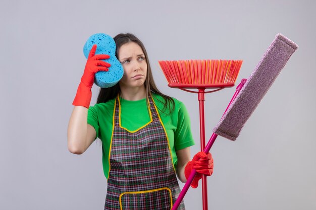 Betrachten der Seite traurige Reinigung junge Frau, die Uniform in den roten Handschuhen hält, die Reinigungswerkzeuge auf isolierter weißer Wand halten