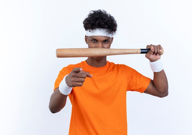 Betrachten des jungen afroamerikanischen sportlichen Mannes der Kamera, der Stirnband und Armband hält, die Fledermaus halten und Sie Geste lokalisiert auf weißem Hintergrund zeigen