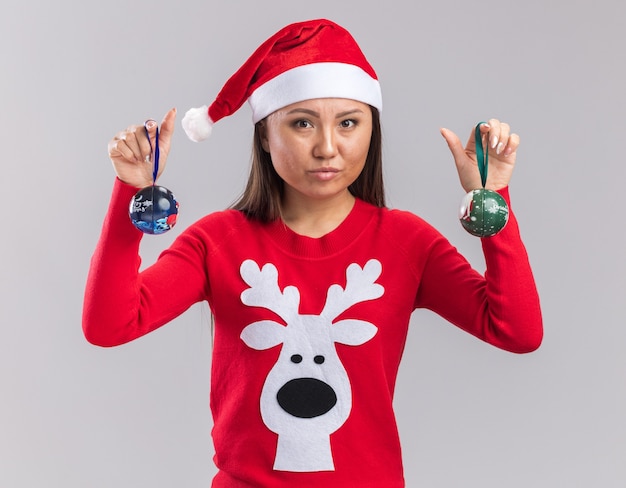 Betrachten des jungen asiatischen Mädchens der Kamera, das Weihnachtshut mit Pullover hält, der Weihnachtsbaumkugeln lokalisiert auf weißem Hintergrund hält