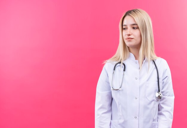 Betrachten des jungen blonden Mädchens des Seitenarztes, das Stethoskop im medizinischen Kleid auf isolierter rosa Wand trägt