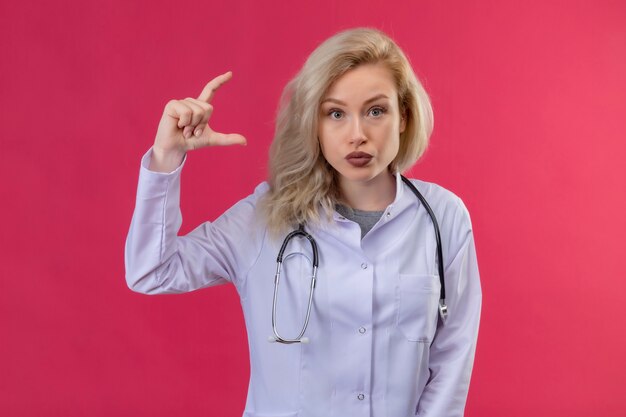 Betrachten des jungen Doktors der Kamera, der Stethoskop im medizinischen Kleid trägt, das Größe auf rotem Hintergrund zeigt
