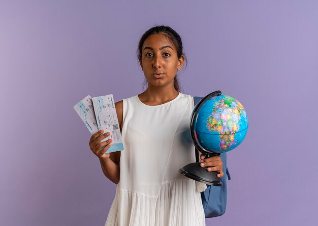 Betrachten des jungen Schulmädchens der Kamera, das Rückentasche hält Globus und Tickets auf lila Hintergrund trägt