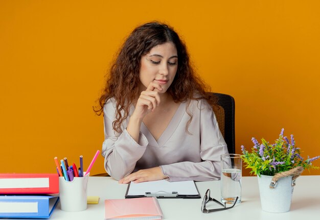 Betrachten des Schreibtischdenkens junge hübsche weibliche Büroangestellte, die am Schreibtisch mit Bürowerkzeugen sitzt, die Hand unter Finger lokalisiert auf orange Wand setzen