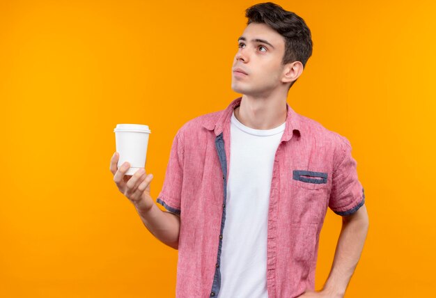 Betrachten des seitlichen kaukasischen jungen Mannes, der rosa Hemd hält, das Tasse Kaffee auf lokalisierter orange Wand hält