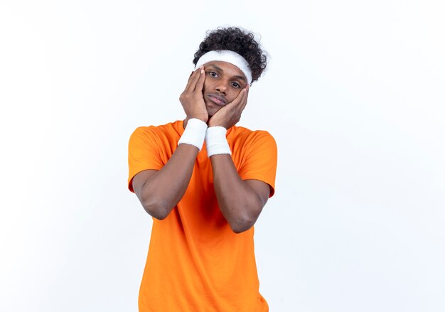 Betrachten des traurigen jungen afroamerikanischen sportlichen Mannes der Kamera, der Stirnband und Armband trägt, die Hände auf Wange lokalisiert auf weißem Hintergrund setzen
