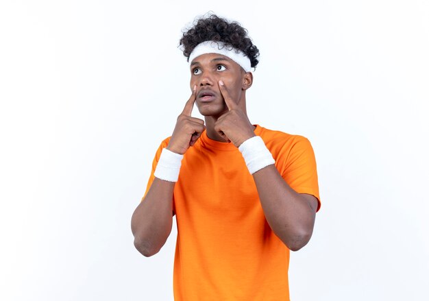 Betrachten des traurigen jungen afroamerikanischen sportlichen Mannes der Seite, der Stirnband und Armbandöffnungsaugen mit den Fingern lokalisiert auf weißem Hintergrund trägt