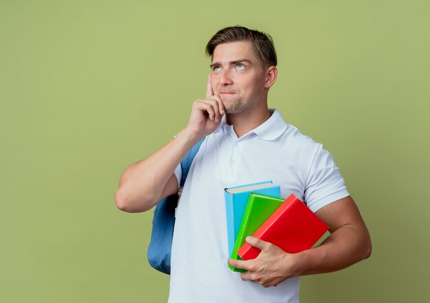 Betrachten nach oben denkender junger hübscher männlicher Student, der Rückentasche hält Bücher hält und Hand auf Wange lokalisiert auf olivgrünem Hintergrund setzt