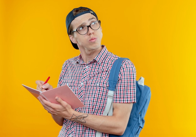 Betrachtet man den denkenden jungen Studentenjungen, der Rucksack und Brille und Kappe hält, die Notizbuch und Stift halten