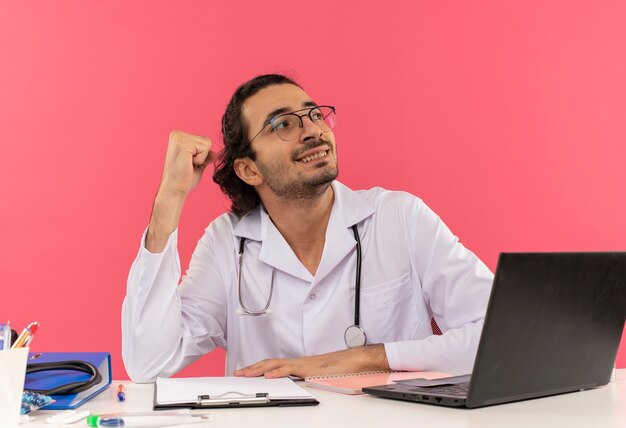Betrachtet man einen lächelnden jungen männlichen Arzt mit medizinischer Brille, der ein medizinisches Gewand mit Stethoskop trägt, das am Schreibtisch sitzt?