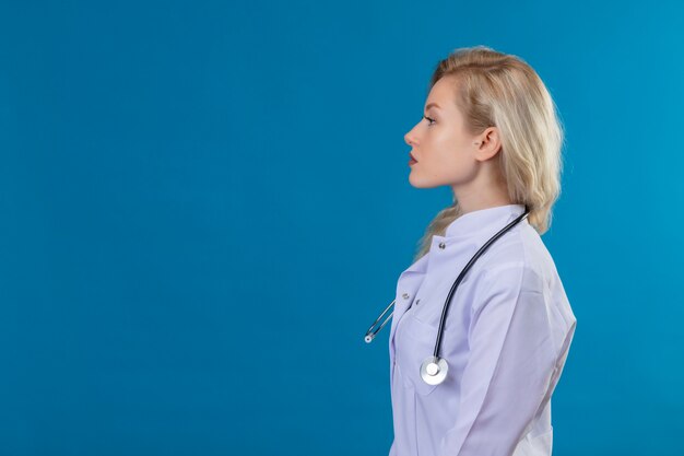 Betrachtet seitlichen jungen Arzt, der Stethoskop im medizinischen Kleid auf blauer Wand trägt