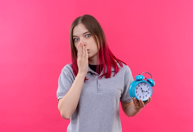 Betroffene junge schöne Frau, die graues T-Shirt hält Wecker hält Mund bedeckt mit Hand auf isolierter rosa Wand