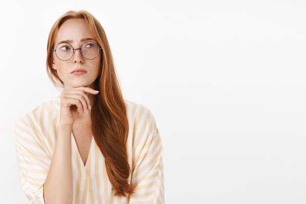 Betroffene nachdenkliche und konzentrierte kreative Schriftstellerin mit roten Haaren und Sommersprossen in Gläsern und trendiger gelber Bluse, die in hmm-Pose steht und rechts zweifelhaft und konzentriert das Kinn berührt