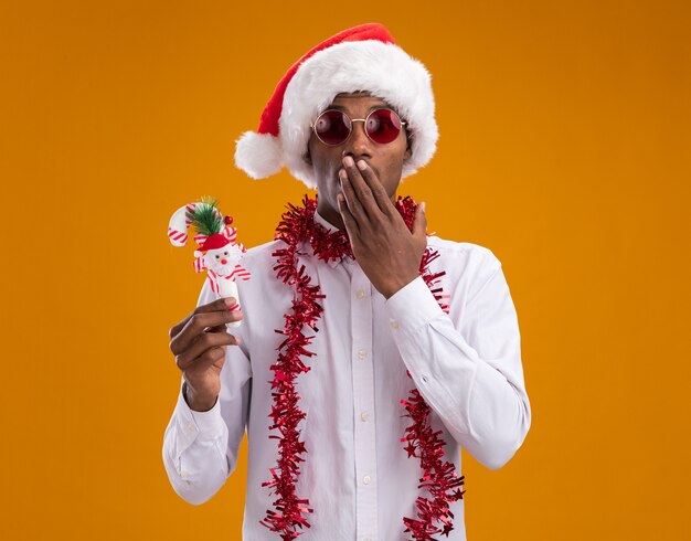 Betroffener junger afroamerikanischer Mann, der Weihnachtsmütze und Brille mit Lametta-Girlande um den Hals hält, der Zuckerstangenverzierung betrachtet, die Kamera betrachtet, die Hand auf Mund lokalisiert auf orangefarbenem Hintergrund hält