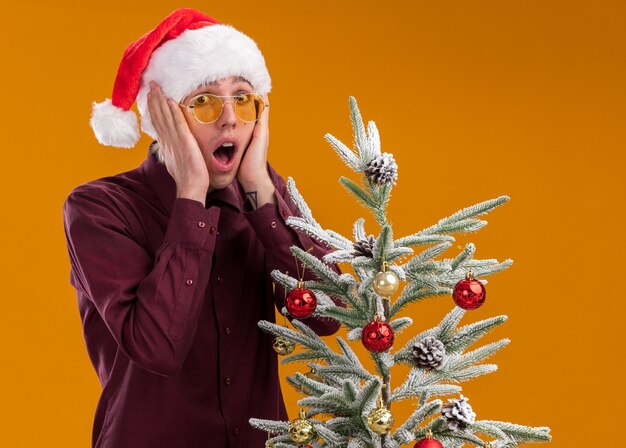 Betroffener junger blonder Mann, der Weihnachtsmütze und Gläser trägt, die nahe geschmücktem Weihnachtsbaum stehen und Hände auf Gesicht lokalisiert auf orange Wand halten