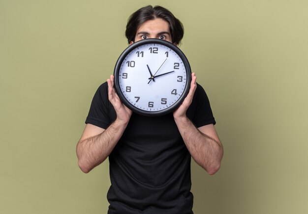 Betroffener junger hübscher Kerl, der schwarzes T-Shirt bedecktes Gesicht mit Wanduhr trägt, die auf olivgrüner Wand lokalisiert wird