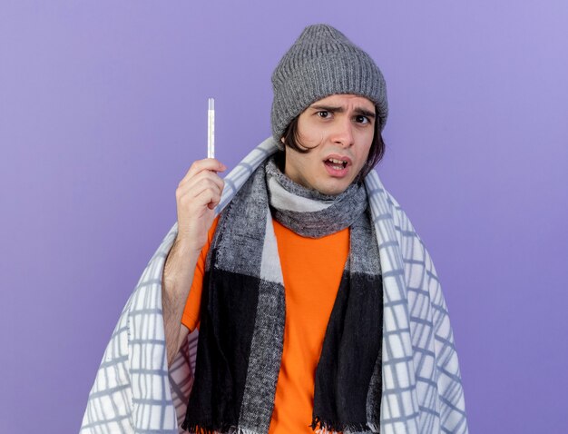 Betroffener junger kranker Mann, der Wintermütze mit Schal wickelt, der im karierten Haltethermometer auf lila Hintergrund eingewickelt ist