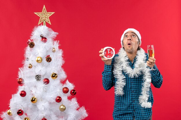 Betroffener junger Mann mit Weihnachtsmannhut und hält ein Glas Wein und eine Uhr in der Nähe des Weihnachtsbaumes